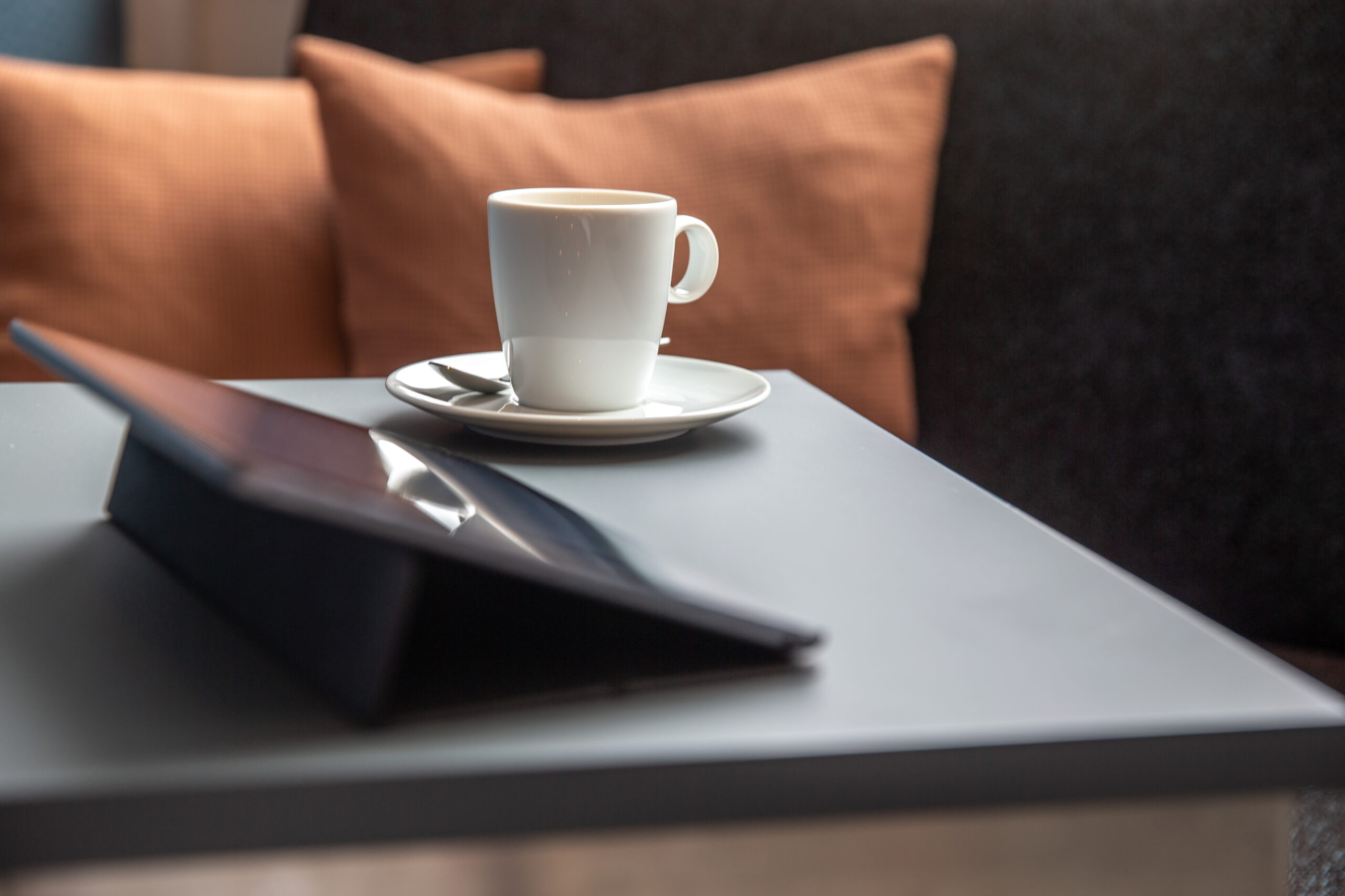 Eine weiße kaffeetasse steht auf einem nidrigen Sofatisch vor einem Ipad. Im Hintergrund sieht man ein oranges Kissen